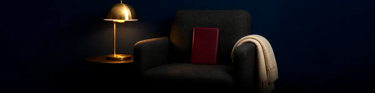 Burgundy leather journal on a charcoal armchair beside a brass lamp in a dim consulting room