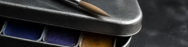 Close-macro of a slightly ajar pewter watercolor tin with unused dry brush bristles on the lid and ultramarine, violet, and ochre pans under cool low-key light
