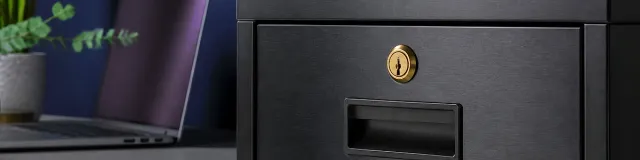 Eye-level close-up of a locked charcoal steel filing cabinet drawer with a brass keyhole; blurred privacy-screen laptop and plant on a cool navy therapy desk