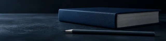 Low side view of a closed untitled navy hardcover and a graphite pencil on a charcoal stone bedside in cool lamp light