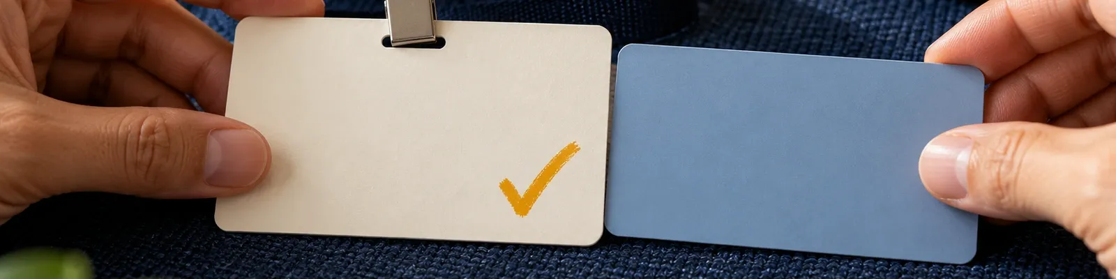 Anonymous hands aligning a blank credential badge and blue network card on navy fabric, with one marigold check mark on the badge.
