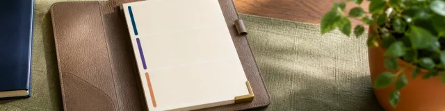 Open taupe padfolio with a blank three-section cream note sheet, navy purple and terracotta bars, on sage linen beside a navy notebook and plant