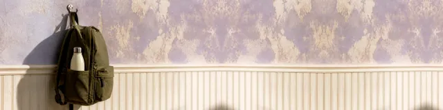 Olive canvas backpack hanging from a small metal wall hook on mottled pale-violet plaster above cream beadboard, closed cream bottle in the side pocket, hard daylight shadow
