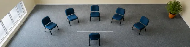 Overhead 4:1 banner of six empty navy clinic chairs in a wide oval on cool slate carpet with a short unlabeled tape line and a sage plant