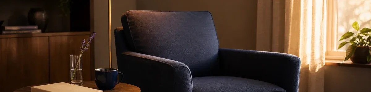 Empty navy armchair beside a closed journal, lavender sprig, and warm reading lamp in a private therapy office