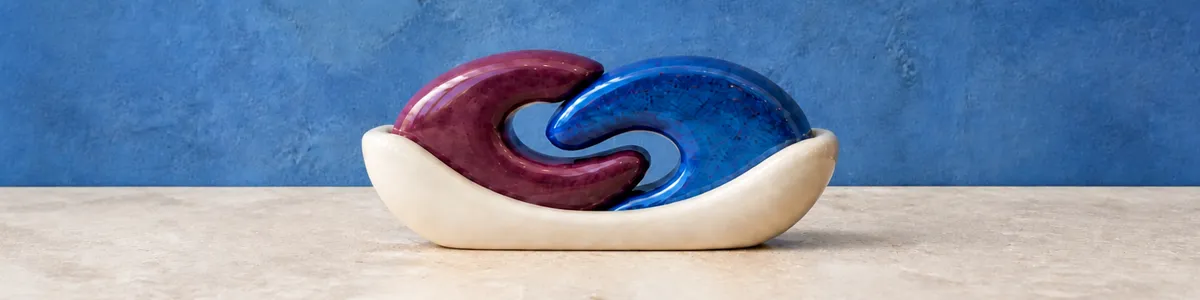 Glossy plum and cobalt curved forms interlock in a cream cradle on a pale stone shelf against a blue wall
