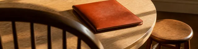 Low view over a dark Windsor chair rail toward a closed terracotta folder on a pale-oak round table in hard late-afternoon raking light, with a second empty chair and a lower stool