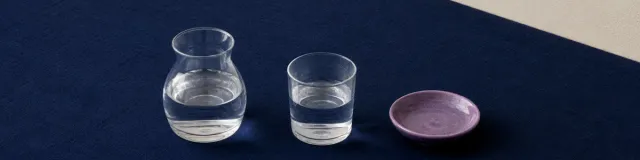 Short clear glass water carafe, matching tumbler, and a muted-purple ceramic dish on a diagonal navy linen cloth against cream plaster