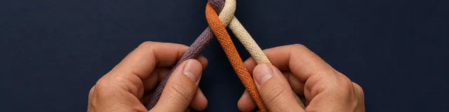 Two hands weaving purple, orange, and cream cords into one braid on a matte navy surface