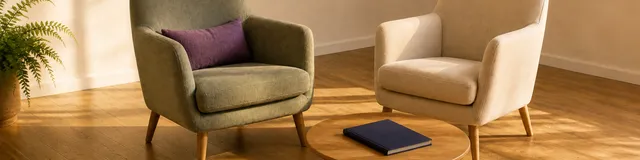 Two empty therapy chairs on oak flooring: sage armchair with a plum cushion, cream chair turned toward it, closed navy notebook on a low oak table
