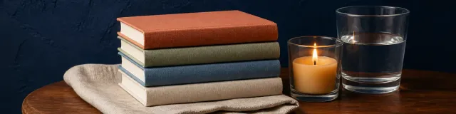 Stack of four blank clothbound notebooks in terracotta, sage, blue, and cream beside a lit candle and water glass on a walnut table against a deep navy wall