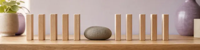 Wooden blocks in a short line on a warm shelf, interrupted by a deliberate gap holding a smooth gray stone, with soft lavender ceramics in the background