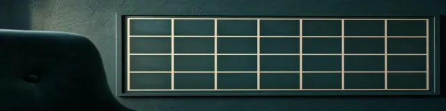 Dark teal tufted chair back beside a framed cream weekly grid on a deep teal plaster wall in soft raking light