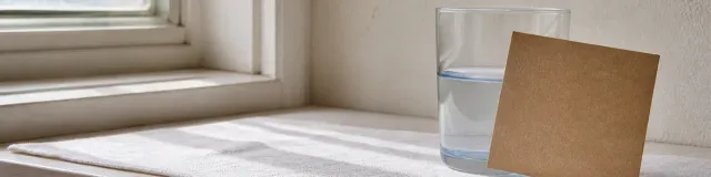 Low three-quarter view of a north-facing windowsill in cool morning side-light, with a clear water glass and one unprinted kraft card leaning against the glass on pale linen