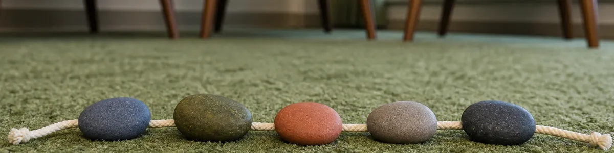 Five smooth stones aligned along a braided cord on a sage rug, with empty recovery-group chairs in the background