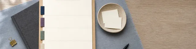 Top-down blue-gray linen desk with wooden clipboard, blank four-band note sheet and navy purple sage sand fabric tabs, charcoal notebook, ceramic dish, brass clip, black pen