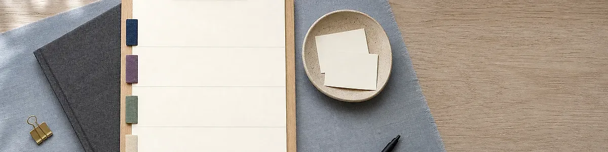 Top-down blue-gray linen desk with wooden clipboard, blank four-band note sheet and navy purple sage sand fabric tabs, charcoal notebook, ceramic dish, brass clip, black pen