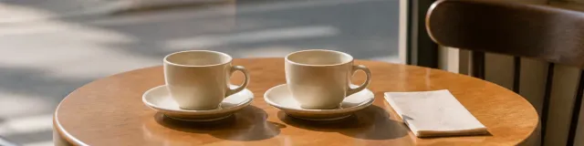 Small round cafe table by a window with two unused ceramic cups on saucers and a folded blank napkin in late-morning light