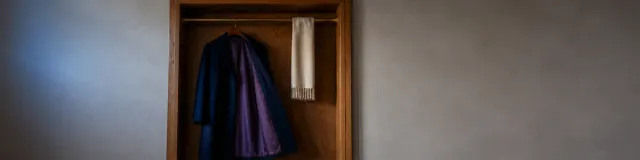 Oak-lined clinic closet with an unused navy wool coat hanging open to show muted-purple lining and a cream scarf on a brass rod, dark-moody plaster to the sides