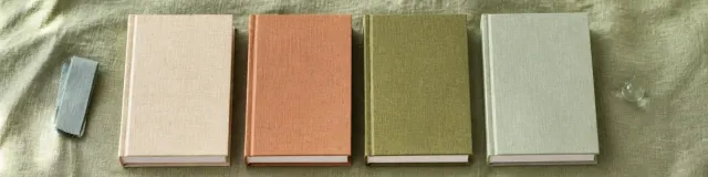Overhead row of four unlabeled cream, terracotta, moss, and celadon cloth workbooks on wrinkled sage linen with a folded cotton tape and a glass pebble