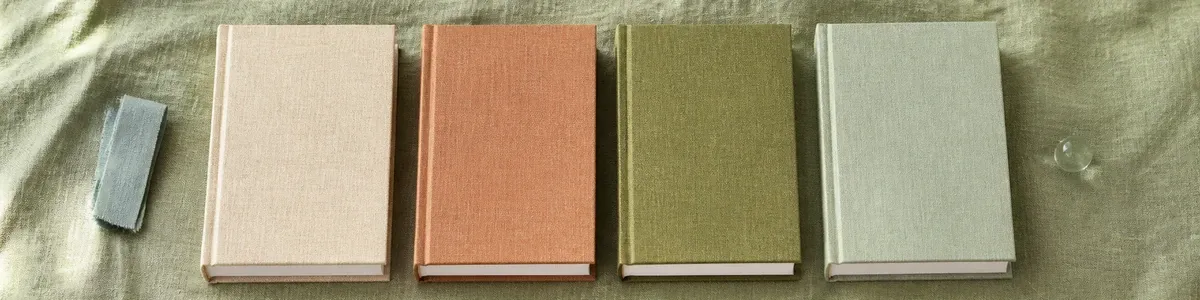 Overhead row of four unlabeled cream, terracotta, moss, and celadon cloth workbooks on wrinkled sage linen with a folded cotton tape and a glass pebble