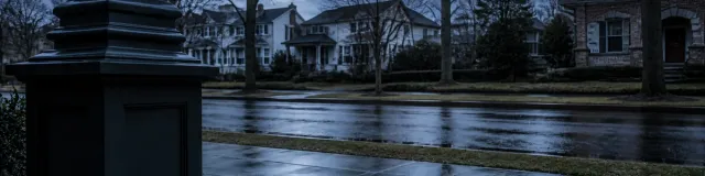 Blue-hour view from a clinic porch past a dark column along a wet slate sidewalk toward an empty residential street