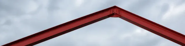 Low upward view of an unused oxide-red steel I-beam gable, both rafters and the bolted ridge plate fully in frame against an overcast gray-blue sky