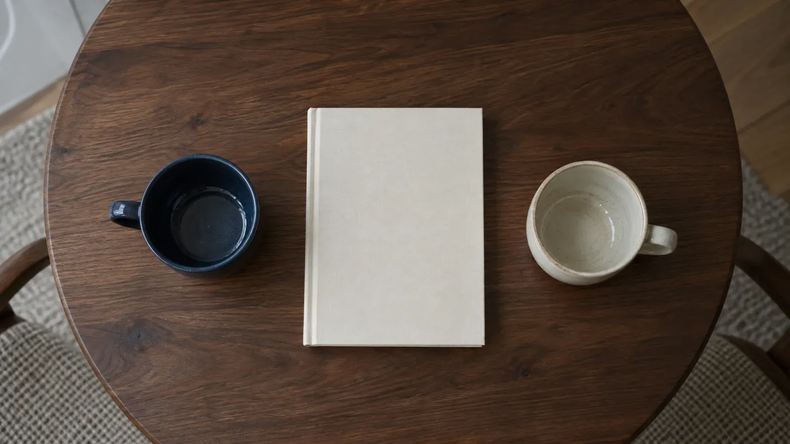Top-down view of a walnut table with a closed cream book between two unmatched cups